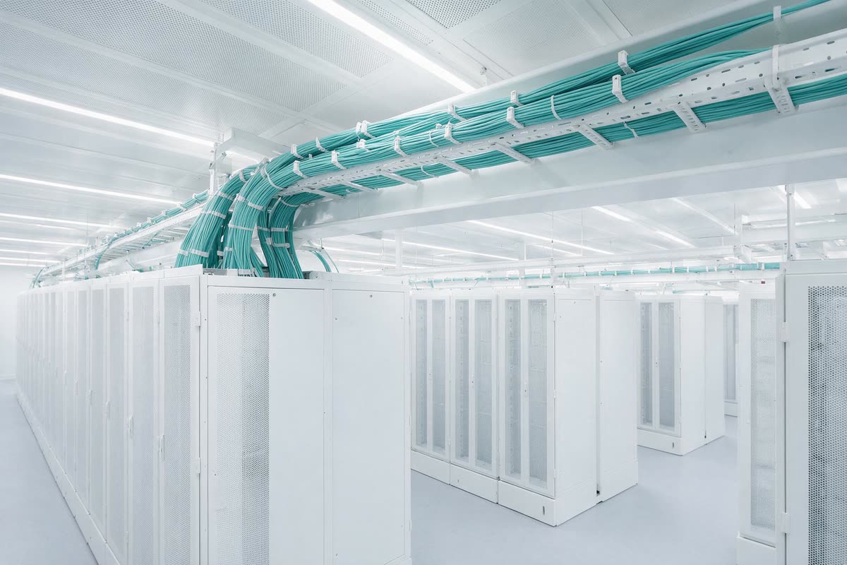 Completed high-density fibre trunking running overhead between rows of white server cabinets in a bright data hall.
